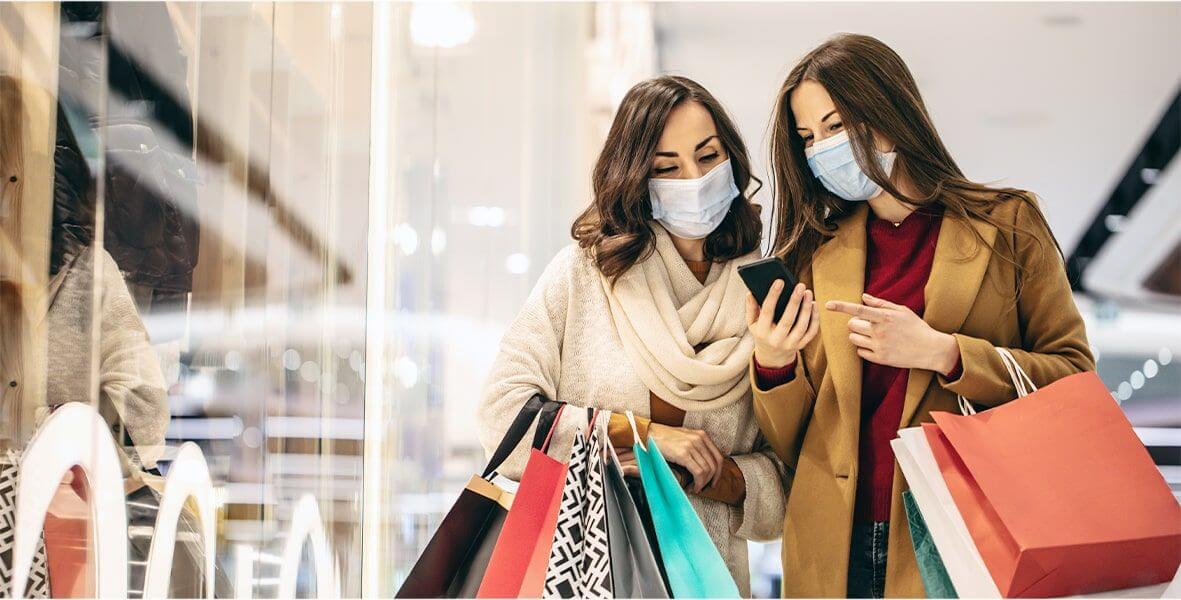 Two women shopping, both wearing masks, carrying bags, and looking at one of their mobile phones.