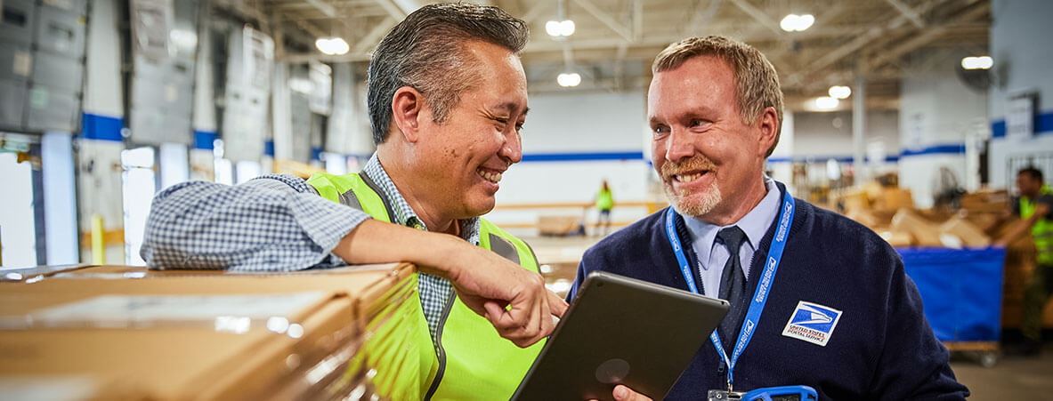 A shipping manager and USPS worker review an order on a tablet over a pallet of packages.
