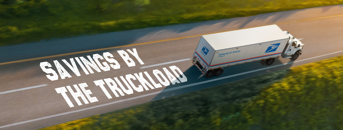 An aerial shot of a USPS delivery vehicle driving down an open road. On the ground, text that reads “SAVINGS BY THE TRUCKLOAD” is left in the wake of the truck.