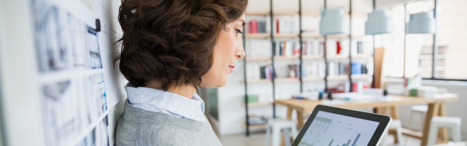 Female professional in an office, leaning against a wall and looking at a tablet.