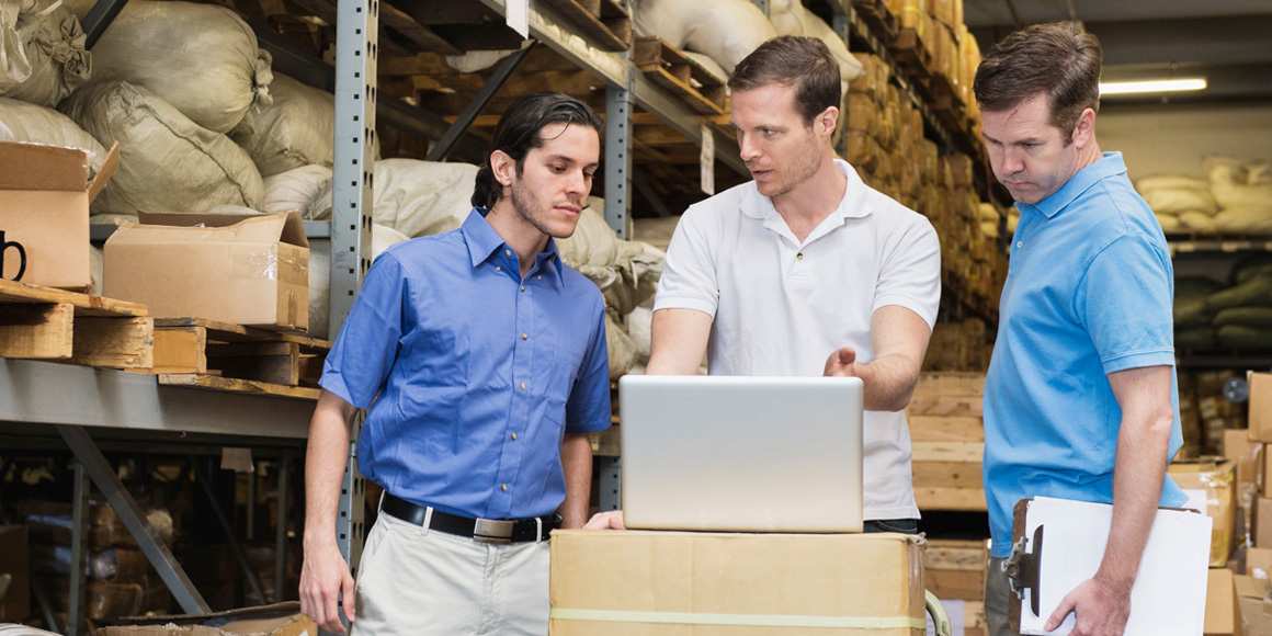 Three warehouse workers in conversation, looking at a laptop.