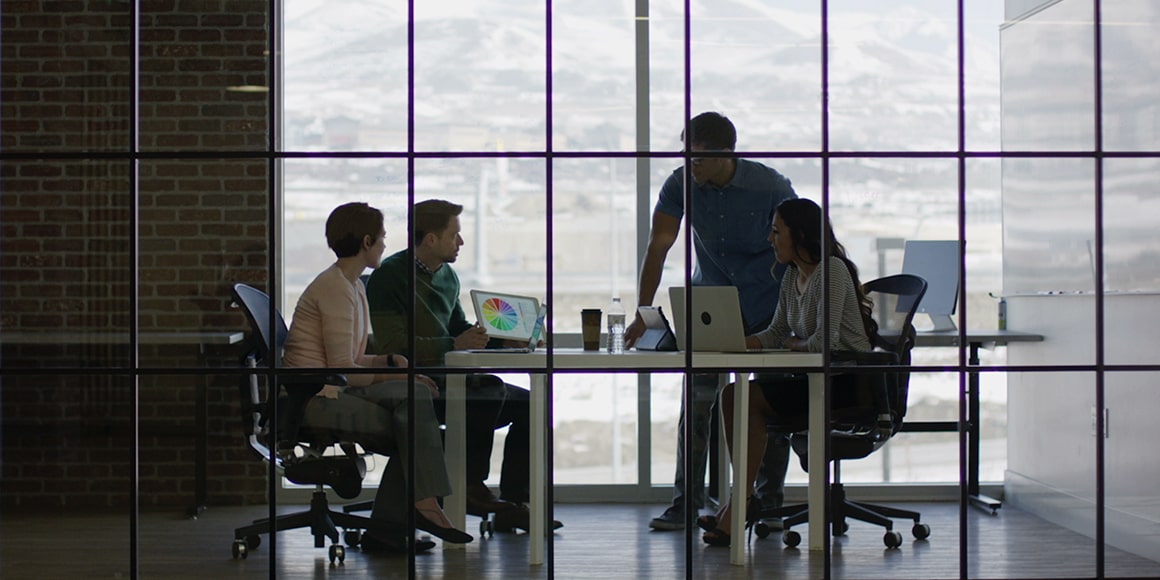 Four people in a glass-enclosed office working together and off computers.