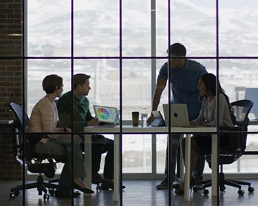 Four people in a glass-enclosed office working together and off computers.