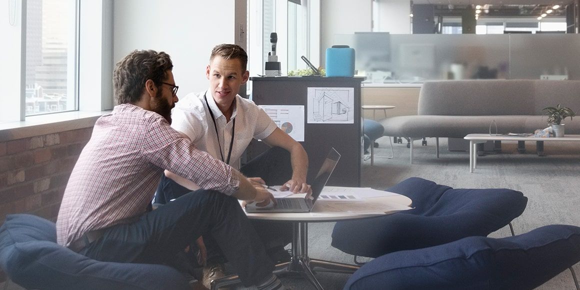 Two male coworkers sitting on lounge chairs in an office, with an open laptop in front of them.