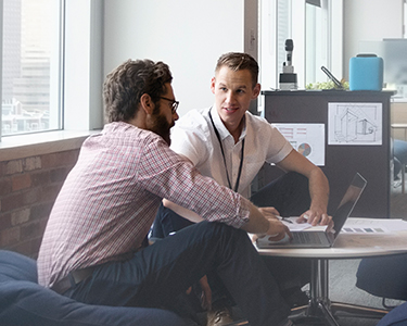 Two male coworkers sitting on lounge chairs in an office, with an open laptop in front of them.