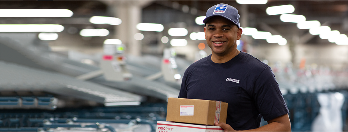 A postal worker at a USPS mail center holding two boxes.