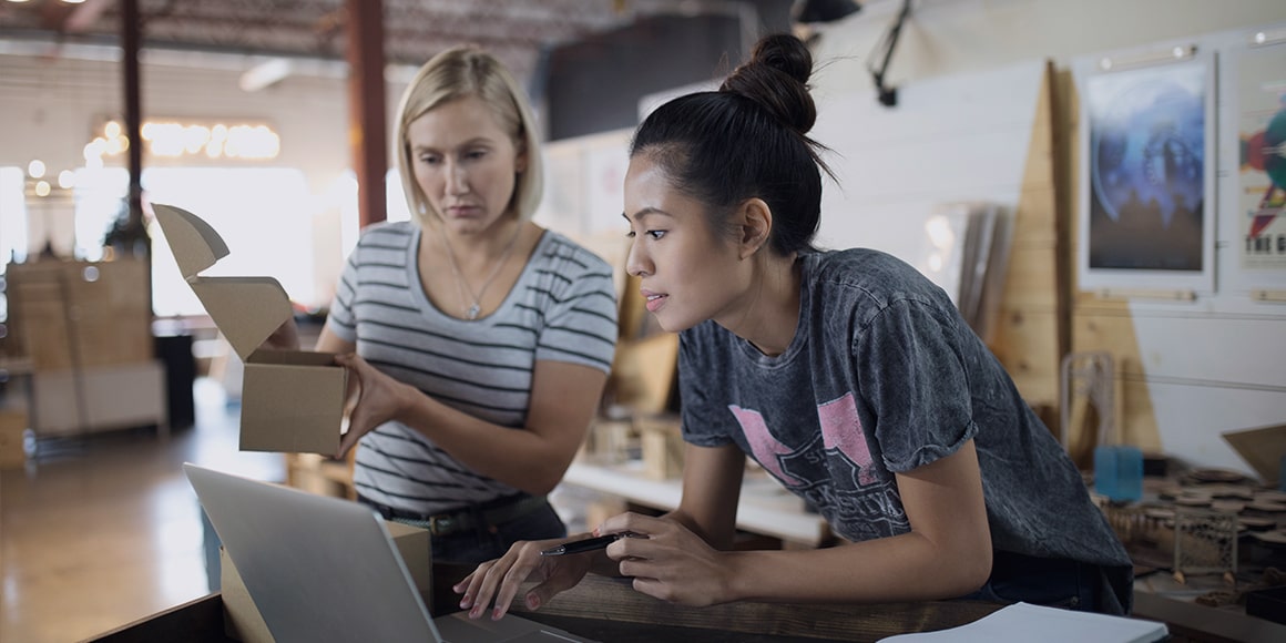 Two women looking at their laptop while one of them is holding a box.