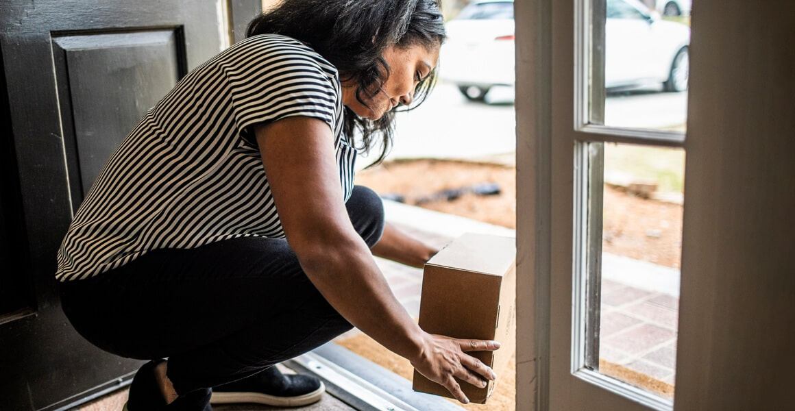 A woman at her doorstep, smiling and kneeling down to look at a package that has been delivered.