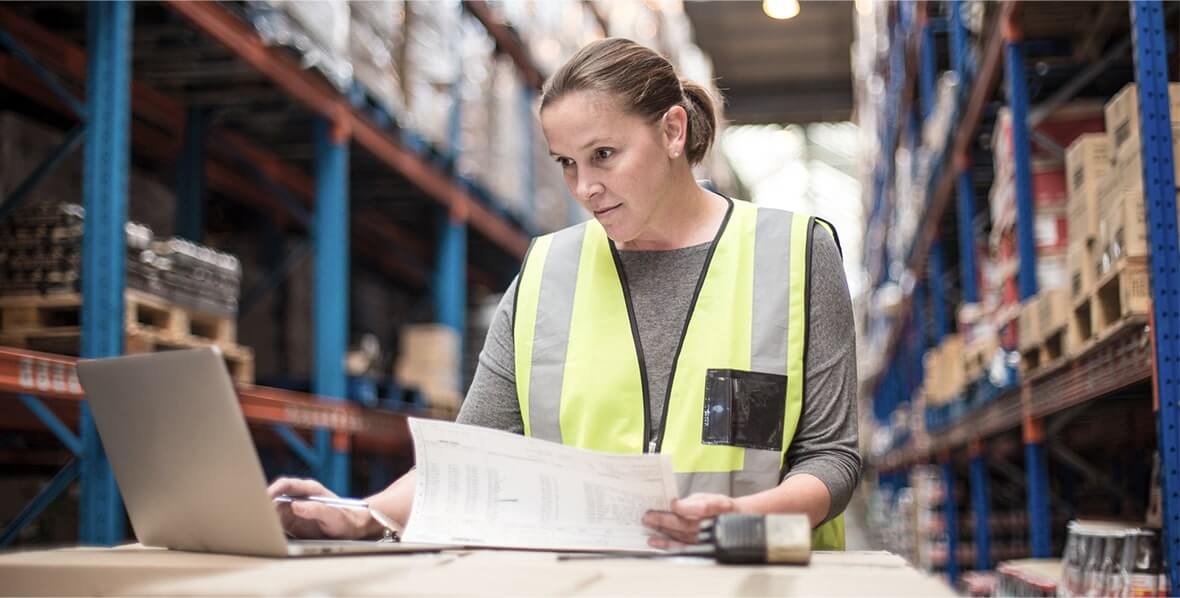 A warehouse worker reading logistics and comparing data against her laptop computer.