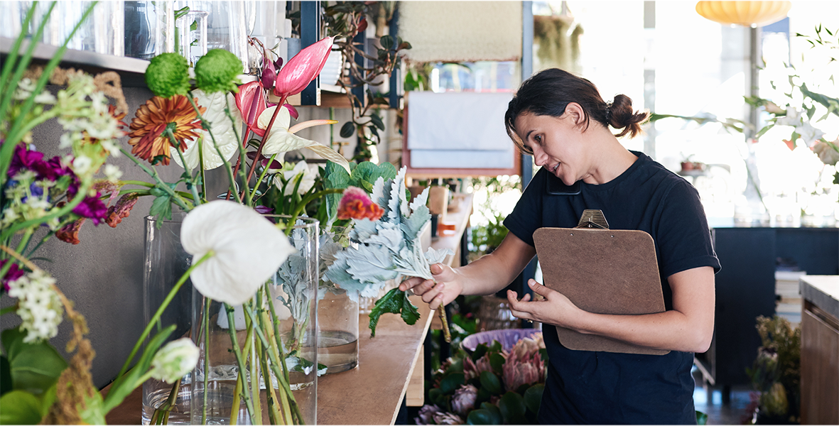 A woman on the phone working in a flower shop looking at a variety of plants.