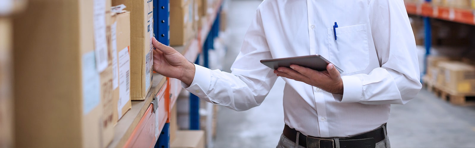 A professional in a white shirt holding a tablet and checking up a package in a warehouse.