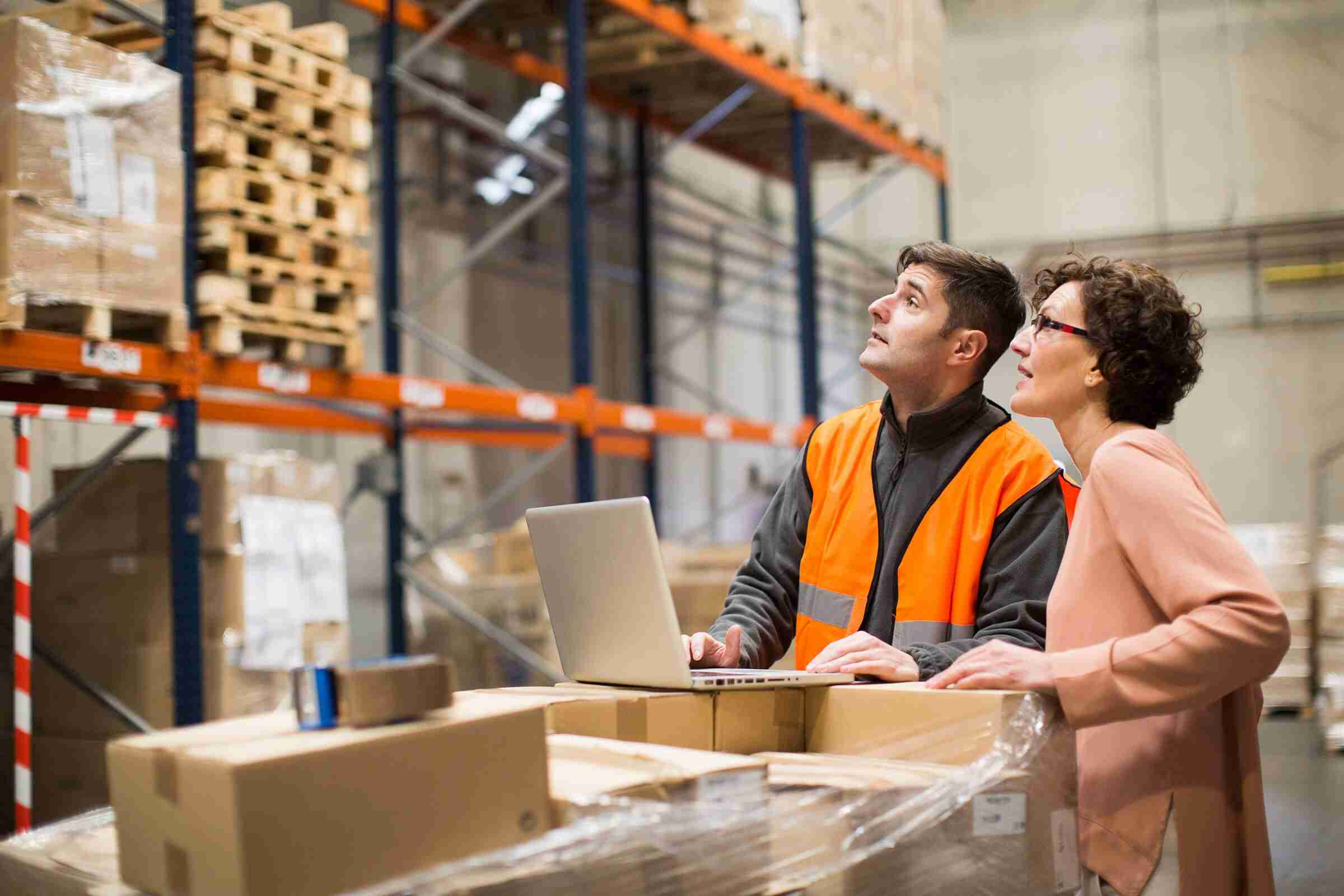 Two warehouse workers with a laptop assessing supplies on warehouse shelves.