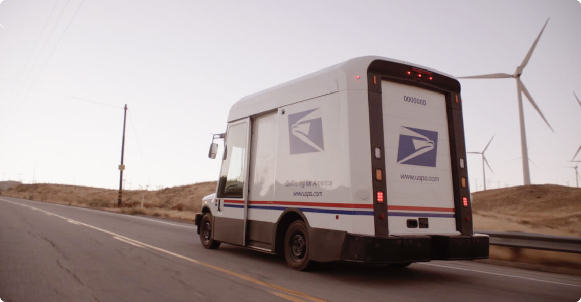 A United States Postal Service Next Generation Delivery Vehicle traveling on a road with wind turbines in the background.